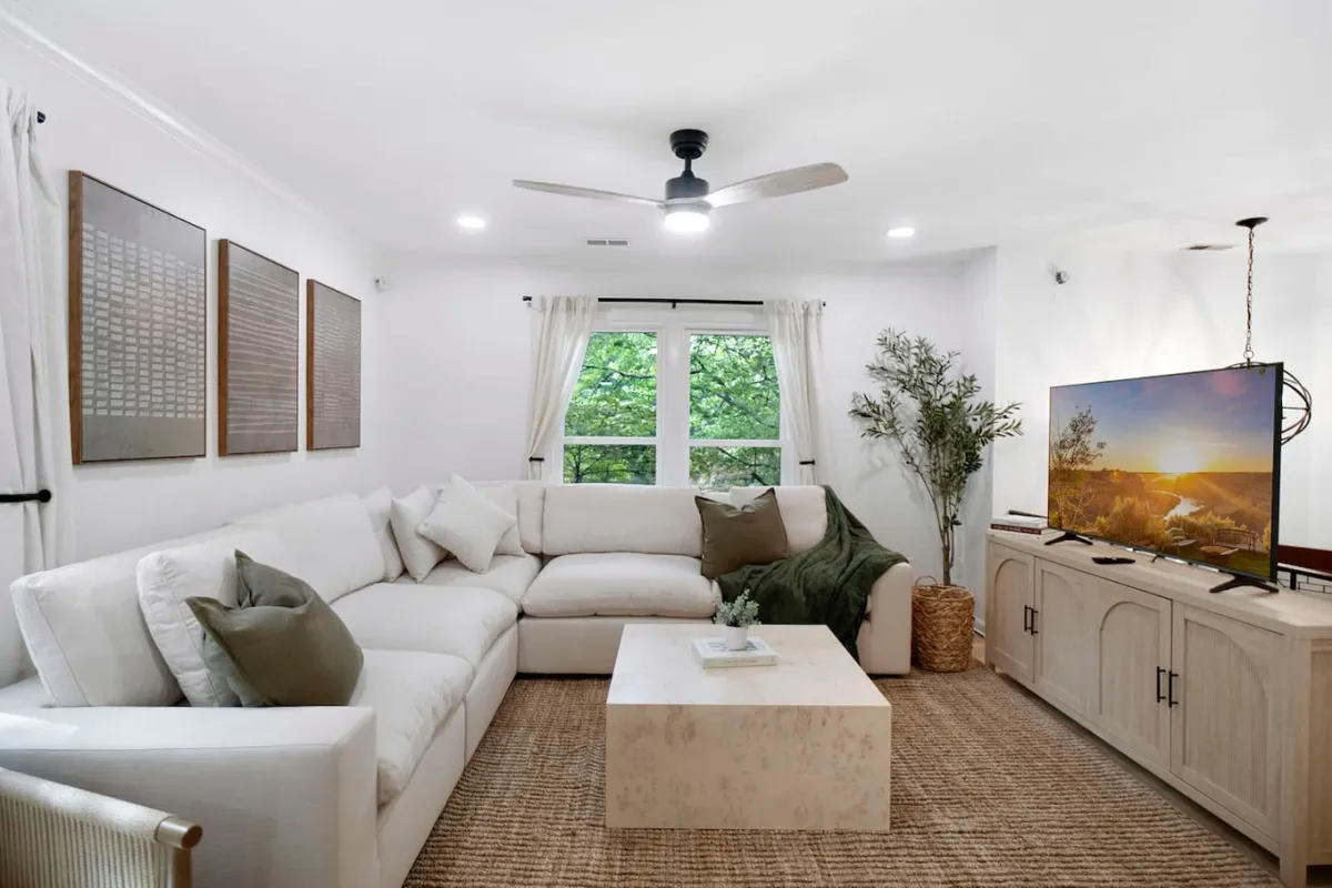 The image showcases a spacious, modern living room with a large, plush sectional sofa arranged around a stylish coffee table and a big-screen television. Natural light pours in through a wide window, revealing inviting views of greenery outside. The room exudes a cozy, welcoming atmosphere, perfect for both quiet relaxation and lively gatherings with family or friends.