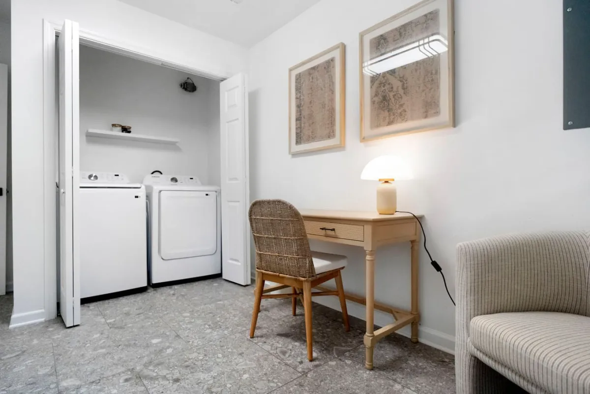 This image presents a practical yet cozy nook featuring a washer and dryer set against a wall, a desk with a lamp, a comfortable chair, and framed artwork. The workspace is adjacent to the laundry facilities, illustrating how guests can multitask between work and daily chores in a single, integrated area.