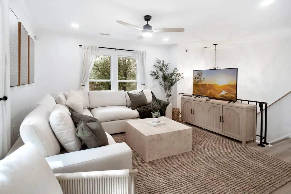 A large white sectional sofa anchors the open lounge, with a coffee table, television, and decorative plant creating a warm, inviting atmosphere perfect for relaxation or movie nights. Sunlight streams through the windows, enhancing the bright and airy mood.