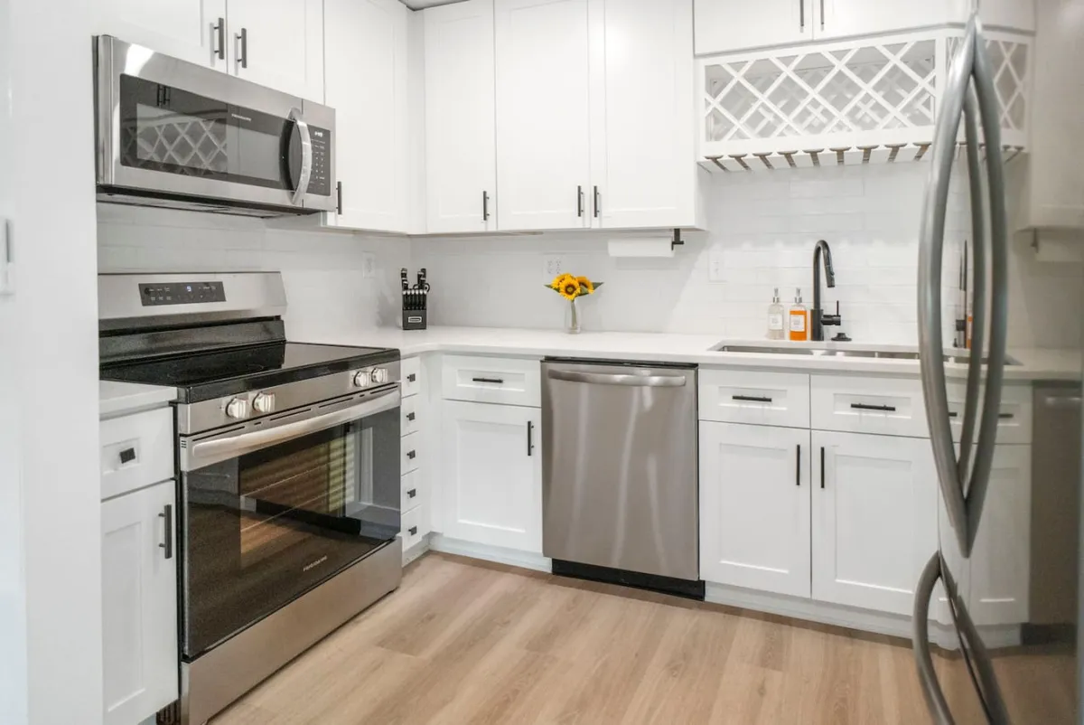 A cheerful kitchen scene featuring stainless steel appliances, white cabinets, sunflowers in a vase, and open shelving. The space is designed for cooking together and entertaining, with plenty of light and room to gather.