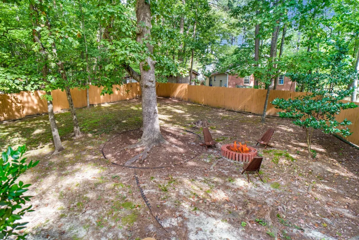 A cozy backyard scene featuring a fire pit surrounded by lawn chairs, bordered by a wooden fence and tall trees. The area invites families to gather for outdoor games, campfires, and picnics, creating a safe and playful environment for children.