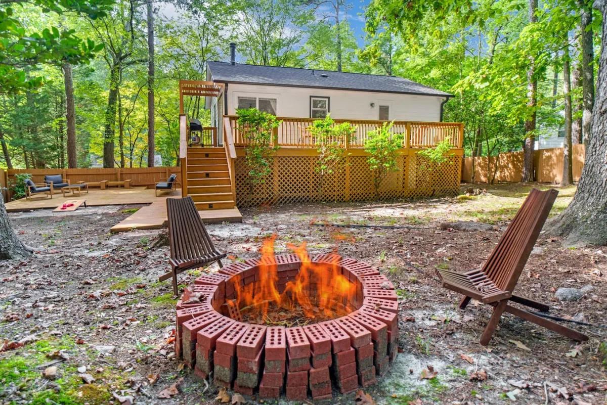 This image captures a wood-burning fire pit surrounded by a cozy seating area on a wooden deck, with a view of the backyard and the house in the background. The scene highlights a relaxed, social atmosphere ideal for gatherings, s’mores, and outdoor relaxation.